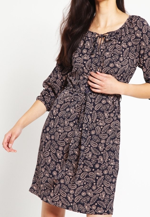 Femme portant une robe fleurie foncée longueur genou, manches trois quarts et un col à cordon, posant avec une main sur la taille.