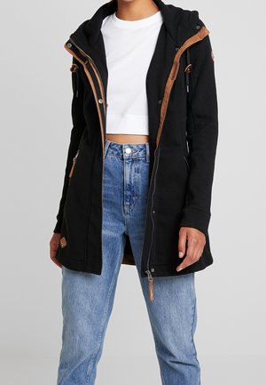 Personne portant une veste à capuche noire avec des garnitures marron sur un top court blanc et un jean bleu taille haute sur un fond uni.