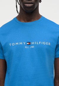 Μπλε βαμβακερό T-shirt με κεντητό λογότυπο "TOMMY HILFIGER" και κείμενο "EST. 1985", στρογγυλή λαιμόκοψη και κοντά μανίκια.