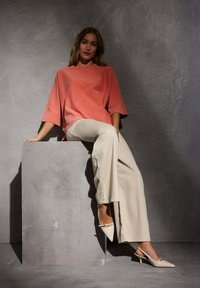 Haut en corail ample à manches courtes, pantalon beige à jambes larges et talons slingback nude. Modèle assis sur un piédestal gris devant un fond texturé.