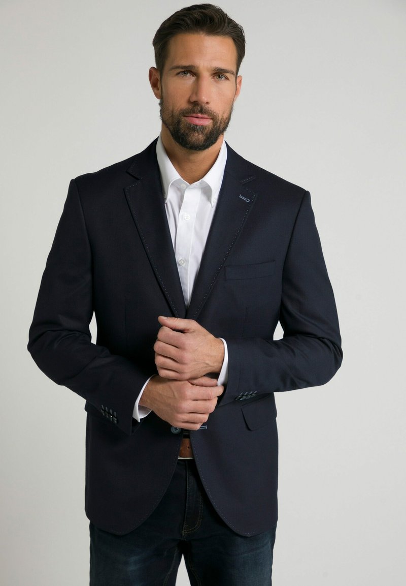 Marinblå blazer med en slät textur, en enda bröstficka och subtila söm detaljer. Kombinerad med en vit skjorta.