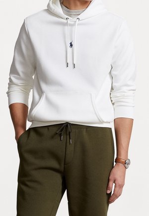 Hombre con sudadera blanca con capucha y logo azul de caballo, pantalones con cordón de color verde oliva, reloj de pulsera en la muñeca izquierda, manos relajadas.