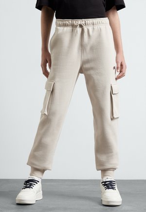 Person iført beige cargobukser med snøre i taljen og hvide sneakers med sorte snørebånd, stående på gråt gulv mod en hvid væg.