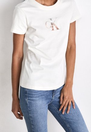 Cream cotton t-shirt with a round neckline and short sleeves, featuring a metallic "ck" logo on the front, paired with blue jeans.