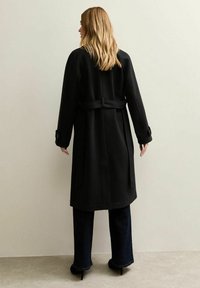 Abrigo negro tipo trench con cinturón en la cintura, corte recto, mangas largas, botones grandes y textura lisa, mostrado desde atrás.