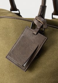 Sac en toile vert olive avec des accents en cuir marron et une étiquette à bagages rectangulaire. L'étiquette présente une finition lisse et une quincaillerie métallique.