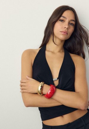 Ung kvinde med langt mørkt hår iført en sort haltertop, røde og guld armbånd, står med krydsede arme mod en ensfarvet baggrund.