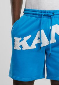 Zilas kokvilnas šorti ar elastīgu jostu un auklu; sānos redzams balts, teksturēts "KAP" logo. Gluda auduma, brīvi pieguļoša piegriezuma.