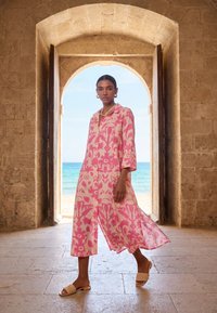 Žena v růžových šatech s květinovým vzorem a sandálech prochází kamennou obloukovou branou s pláží a mořem v pozadí.