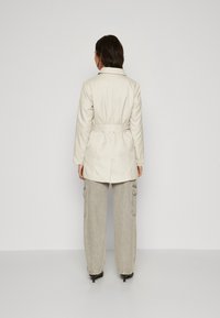 Geacă lungă bej cu talie betelată, având un guler clasic și o siluetă dreaptă, purtată peste pantaloni cargo gri. Textură netedă.