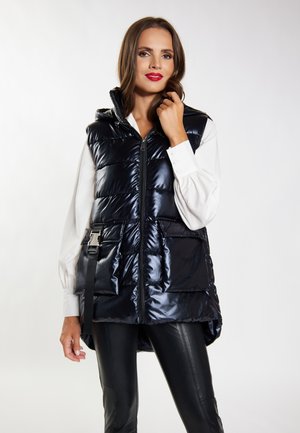 Zwarte puffervest met een glanzende afwerking, ritssluiting, capuchon en twee voorzakken. Draagt over een witte blouse met lange mouwen en zwarte broek.