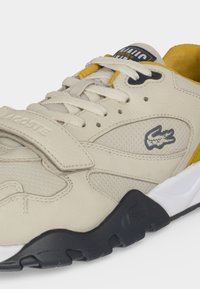 Μπεζ αθλητικό Lacoste με λευκά κορδόνια, κίτρινη εσωτερική επένδυση, μαύρη και λευκή σόλα και λογότυπο κροκόδειλου στο πλάι.