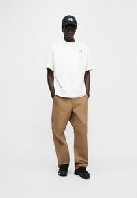 Λευκό βαμβακερό t-shirt, άνετη εφαρμογή, μικρό λογότυπο στο στήθος· καφέ παντελόνι cargo με τσέπες, μαύρα αθλητικά παπούτσια, μπλε σκουφάκι με λογότυπο.