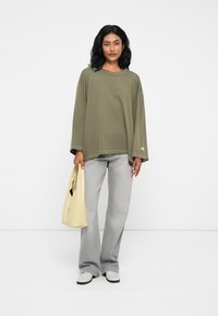 Υπερμεγέθης μπλούζα ελιάς με μακριά μανίκια, ανοιχτό γκρι φαρδύ παντελόνι, κρεμ παπούτσια, κρατώντας μια κίτρινη τσάντα tote με κοψίματα.