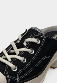 Μαύρο καμβαδένιο παπούτσι με τακούνι, χωρίς κορδόνια, με άσπρο ραφές, μεταλλικές τρύπες για κορδόνια και χοντρά, εκρού κορδόνια δεμένα σε χαλαρό φιόγκο.