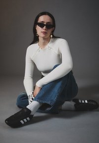 Weißes Langarmoberteil mit Ringdetails, blaue weit geschnittene Denim-Jeans, schwarze Sneakers mit weißen Streifen und Sonnenbrille. Schlichter Hintergrund.