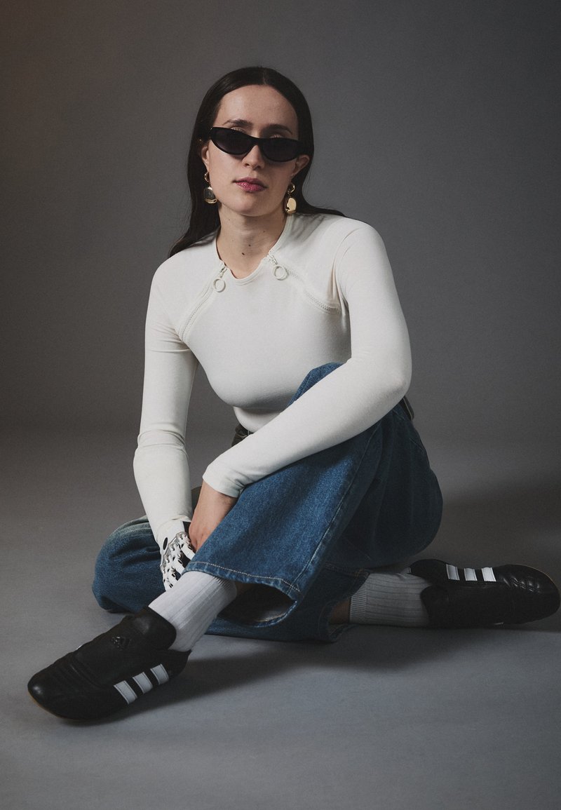Weißes Langarmoberteil mit Ringdetails, blaue weit geschnittene Denim-Jeans, schwarze Sneakers mit weißen Streifen und Sonnenbrille. Schlichter Hintergrund.