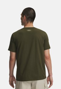 Camiseta de algodón verde oliva con mangas cortas, escote redondo y un pequeño logo en un tono más claro justo encima del cuello en la parte posterior.