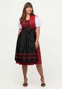 Červený a bílý dirndl s květinovým vzorem, černý saténový zástěr, šněrovací korzet a krátké nabírané rukávy; délka ke kolenům s stužkovými akcenty.