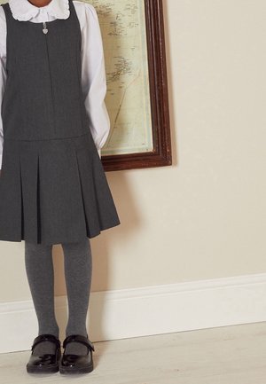 Bambina che indossa un grembiule grigio plissettato, una camicetta bianca con colletto arricciato, calze grigie e scarpe Mary Jane nere, in piedi vicino a un muro con una mappa incorniciata.