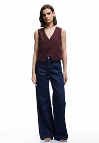 Gilet sans manches bordeaux avec un col en V et deux poches avant, associé à un jean en denim noir à jambes larges. Détail de fermeture à boutons simple.