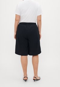 Zwarte, knielange shorts met een elastische tailleband. De stof lijkt lichtgewicht. Er wordt een wit T-shirt gedragen en de focus ligt op de achterkant van de shorts.