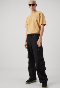 Gul kortermet T-skjorte med logo, kombinert med svarte cargo-bukser med flere lommer og svarte sneakers med grønne detaljer.