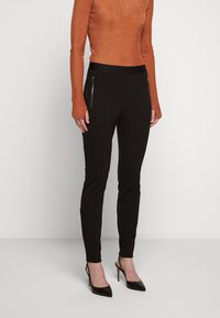 Pantaloni neri aderenti con tasche laterali con zip, abbinati a un maglione arancione a coste. I pantaloni hanno una texture liscia e un design affusolato.