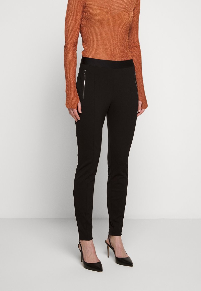 Pantaloni neri aderenti con tasche laterali con zip, abbinati a un maglione arancione a coste. I pantaloni hanno una texture liscia e un design affusolato.