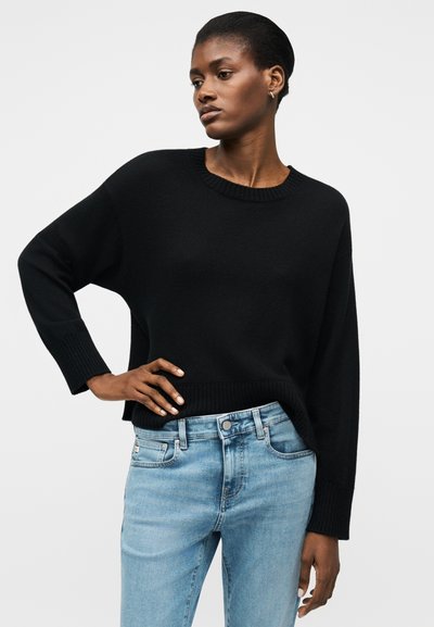 Η γυναίκα με κοντά μαλλιά φοράει ένα μαύρο πουλόβερ με μακριά μανίκια και ανοιχτό μπλε τζιν, στέκεται με το ένα χέρι στη μέση, μπροστά από απλό φόντο.