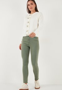 Cárdigan blanco de punto con cuello redondo, botones de perla y detalles verdes, combinado con jeans skinny verdes y zapatillas blancas.