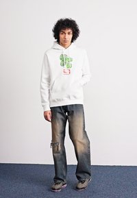 Weißer Hoodie mit grünem Pflanzenmotiv und rotem Etikett, kombiniert mit ausgebleichten Jeans mit einem aufgerissenen Knie und grauen Turnschuhen.