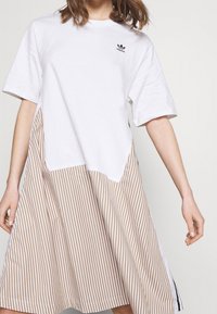 Valkoinen T-paitamekko, jossa on raidallinen beige ja valkoinen epäsymmetrinen helma. Mekossa on lyhyet hihat ja Adidas-logot rinnassa.
