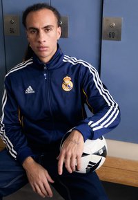 Ensemble de survêtement zippé bleu marine avec des bandes blanches Adidas et le logo du Real Madrid. Le mannequin tient un ballon de football noir et blanc, assis sur un banc en bois.