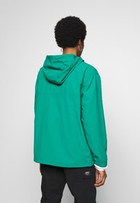 Grøn hættetrøjejakke med letvægtsmateriale, elastiske manchetter og et enkelt design. Har en synlig søm tværs over ryggen.