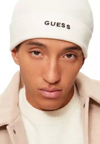 Beanie bianco con logo "GUESS" nero, texture morbida in maglia. Modello che indossa un maglione color crema e un soprabito beige chiaro. Sfondo neutro.