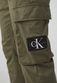 Olivově zelené cargo kalhoty se boční kapsou a černou nášivkou s nápisem "CK" a "Calvin Klein Jeans" v bílém písmu. Textura bavlněné tkaniny.