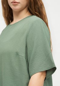 Γυναίκα με μακριά κόκκινα μαλλιά που φοράει ένα υφασμάτινο πράσινο-φασκόμηλο κοντομάνικο τοπ σε απλό ανοιχτό φόντο.