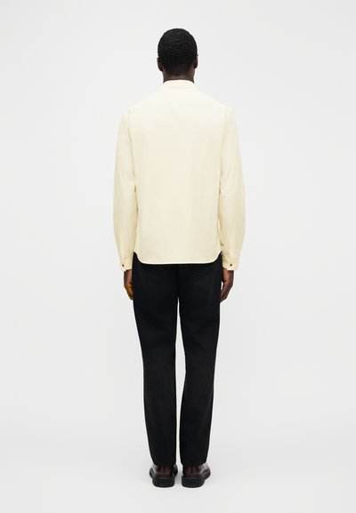 Jasnożółta koszula z długim rękawem o zaokrąglonym dole, noszona z czarnymi spodniami i ciemnymi butami, widziana od tyłu. Gładka faktura, minimalne detale.