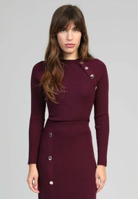 Femme aux longs cheveux bruns portant une robe bordeaux côtelée ajustée avec des boutons dorés décoratifs sur l'épaule et la jupe, se tenant contre un fond gris.