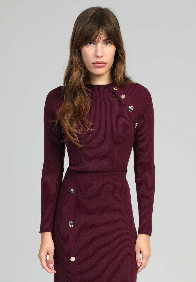 Femme aux longs cheveux bruns portant une robe bordeaux côtelée ajustée avec des boutons dorés décoratifs sur l'épaule et la jupe, se tenant contre un fond gris.