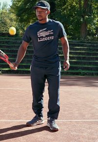 Camisa de manga curta azul-marinho com texto gráfico, calças escuras e sapatilhas de treino cinzentas. A segurar uma raquete de ténis num campo de terra batida rodeado de vegetação.