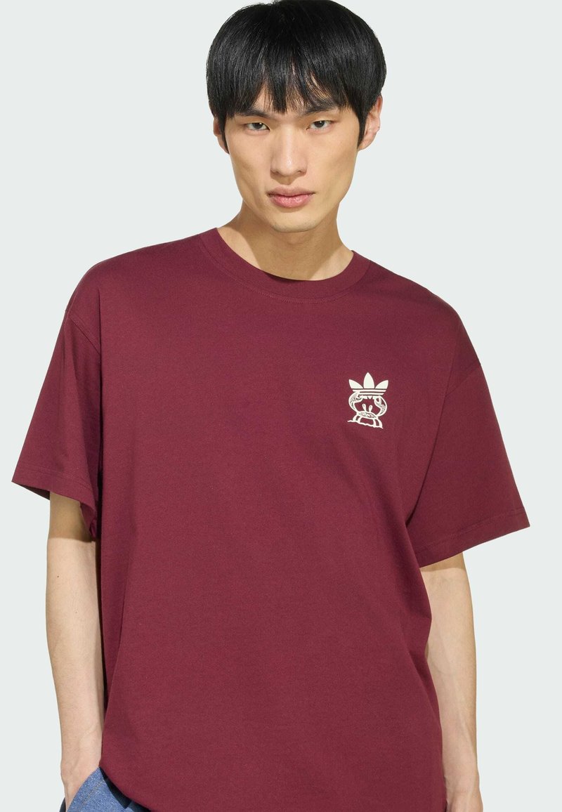 Junger Mann mit glatten schwarzen Haaren, der ein kastanienbraunes T-Shirt mit einem kleinen weißen Grafiklogo auf der linken Brust trägt.