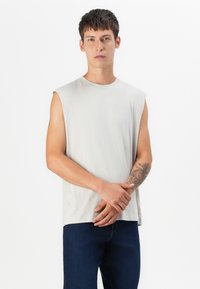 Camiseta sin mangas de un tono gris claro, fabricada en un tejido suave, con un escote redondo y un corte relajado. El modelo lleva unos pantalones vaqueros oscuros y tiene un tatuaje en el brazo izquierdo.