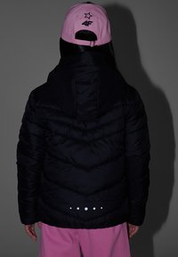 Personne de dos portant une doudoune noire, une casquette de baseball rose avec une étoile et le logo 4F, et un pantalon rose ample sur un fond gris.