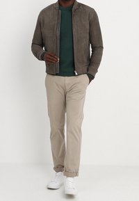 Giubbotto bomber in suede grigio su un maglione a coste verde, abbinato a pantaloni beige e sneakers bianche, con chiusura a zip e polsini arrotolati.