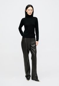 Maglione nero a collo alto realizzato in materiale morbido, abbinato a pantaloni in pelle neri a vita alta. Scarpe nere con punta e dettagli dorati.