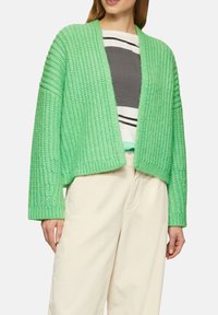 Grüne Strickjacke mit offenem Vorderteil, gerippter Textur und langen Ärmeln. Getragen über einem grau-weißen gestreiften Oberteil und hellen Hosen.