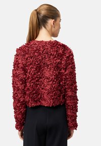 Veste courte rouge à texture florale avec un design de fleurs en 3D, dotée de manches longues et d'un col rond, associée à un pantalon noir.