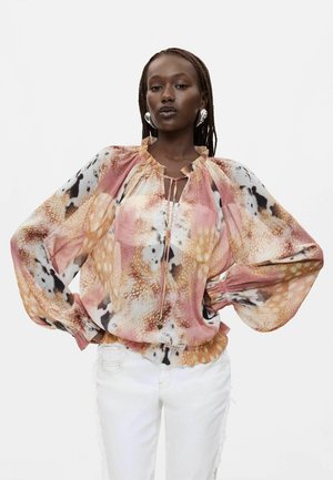 Vrouw met gevlochten haar die grote zilveren oorbellen draagt en een losse, meerkleurige blouse met ballonmouwen en witte broek.